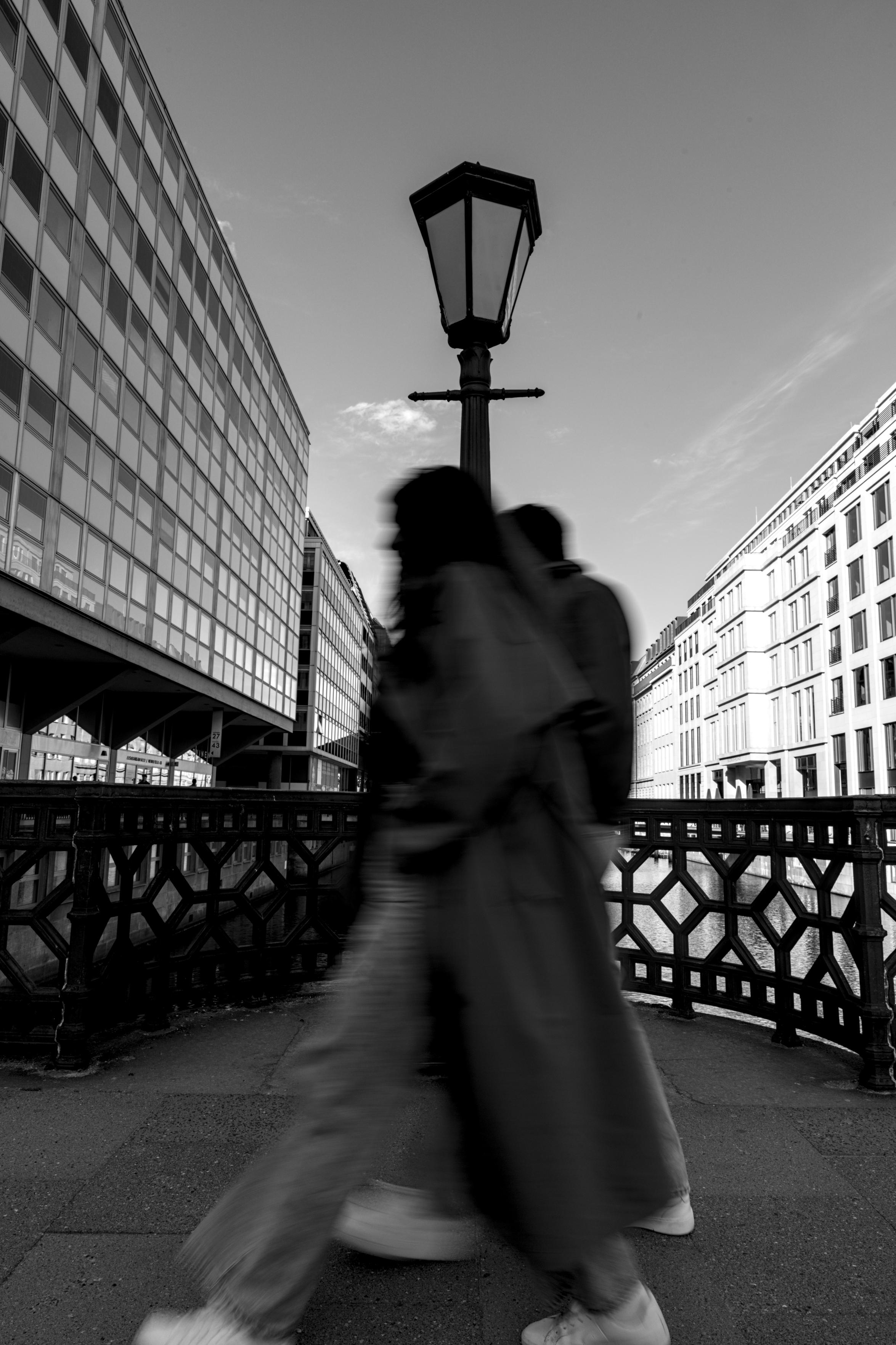 Zwei leicht verschwommene Personen gehen durchs Bild, direkt hinter ihnen steht eine Laterne. Links und Rechts Häuserfassaden.