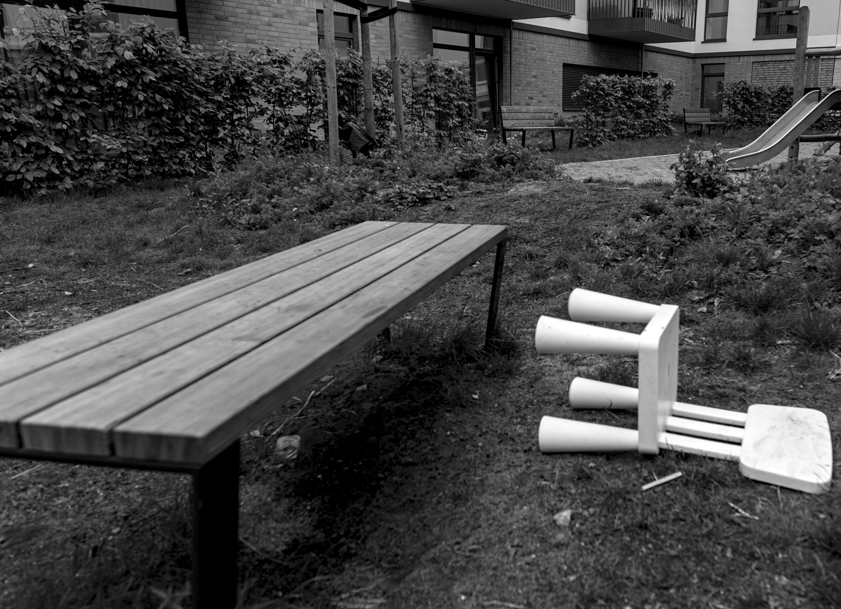Schwarzweiß Bild. Links steht eine Holzbank, rechts davor ein umgefallener, weißer Plastikstuhl auf einem Rasen. Im Hintergrund eine Rutsche und die Hausfassade.