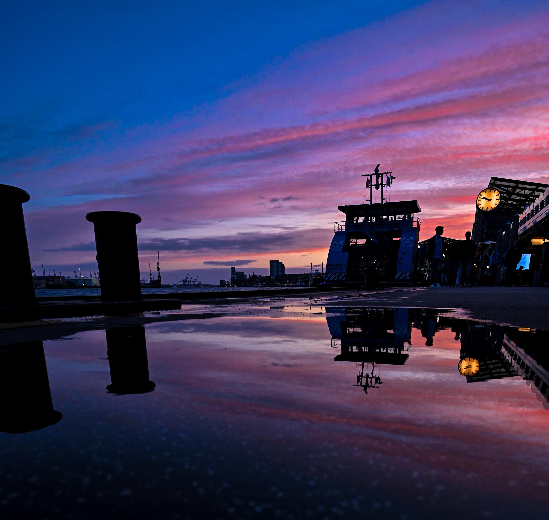 Sonnenuntergang, blauroter Himmel, am Ponton liegt eine Fähre, die sich in einer Pfütze auf dem Ponton spiegelt.