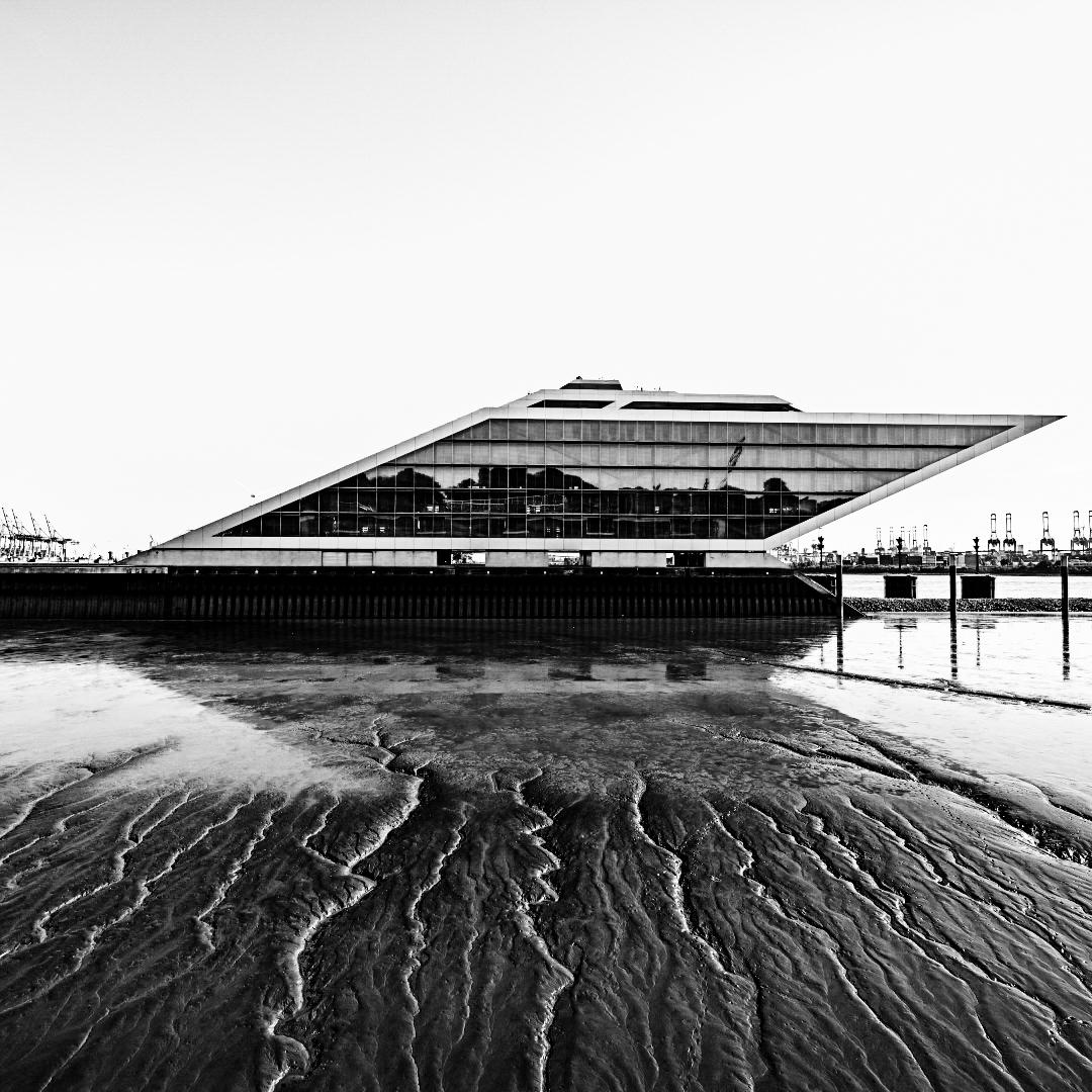 Das Dockland ( ein Gebäude in einer nach vorn geneigten Form) von der Seite fotografiert, davor der Elbschlick bei Ebbe mit sein vertikal verlaufenden Rinnsalen