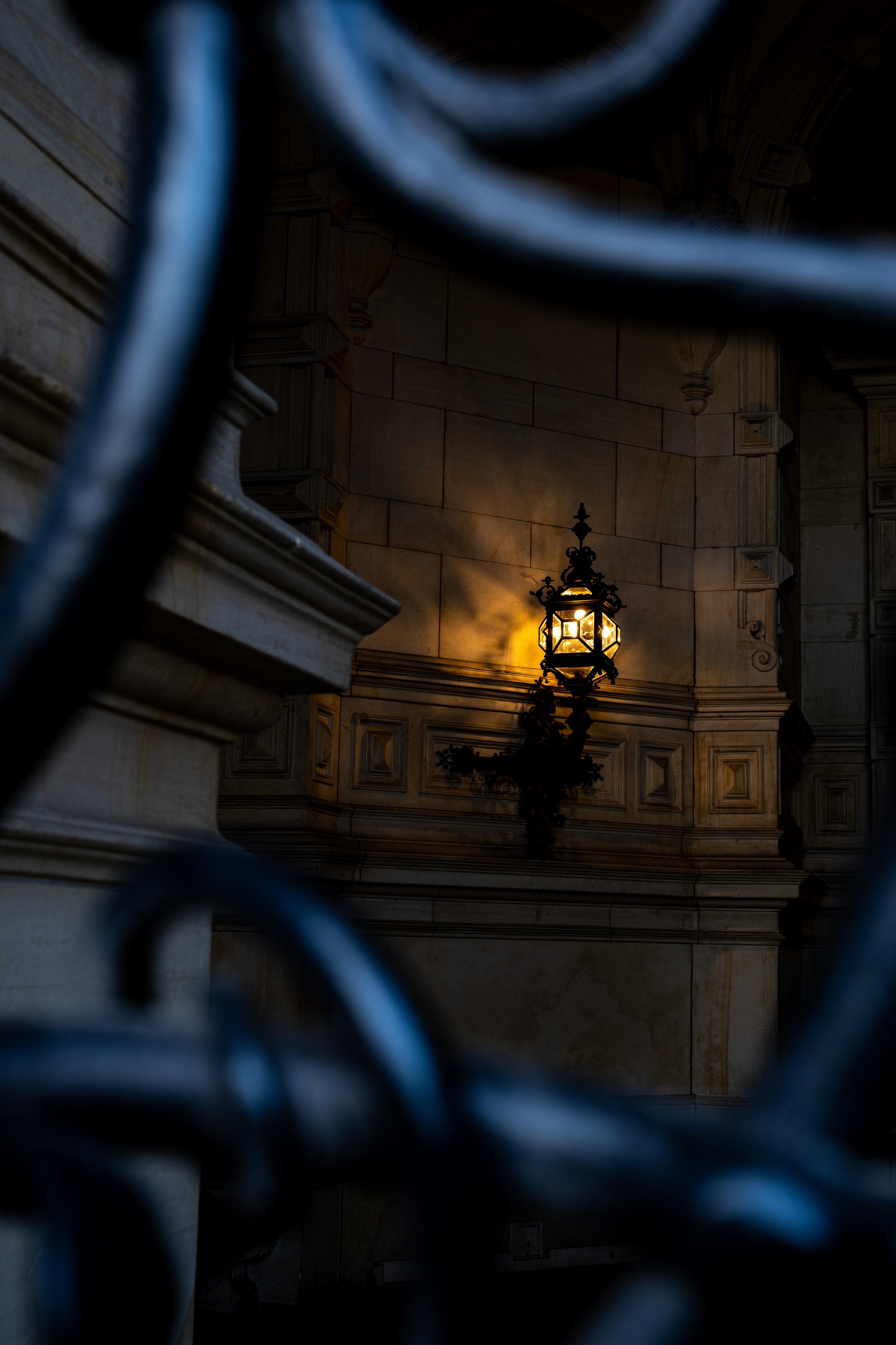 Durch ein verziertes Torgitter eine alte verzierte Wandlampe die leuchtet fotografiert. Die Lampe hängt an einer Wand verziert mit einer gemauerten Bordüre.